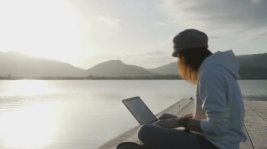 İnternet serbest meslek seçimi konsepti: genç bir kadın gün batımında bir gölün kenarında dizüstü bilgisayarıyla çalışıyor.