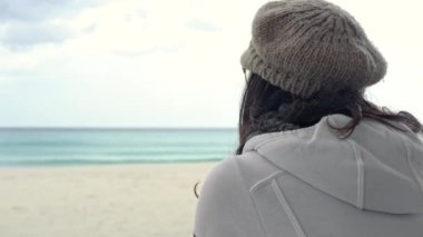 Kışın fırtınalı bir gökyüzü ile deniz kenarındaki kapalı bir barın güneş odasındaki tahta direğe yaslanmış genç bir kadın dalgın ufka bakar.