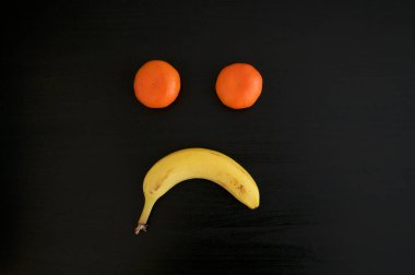 Mandalinalardan ve siyah arka planda muzdan yapılmış hüzünlü bir somurtma. Bu kavramsal fotoğraf için üzüntü, depresyon ve olumsuzluk olarak kullanılabilir.