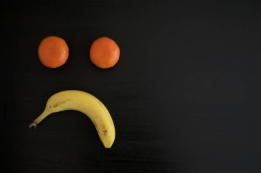Mandalinalardan ve siyah arka planda muzdan yapılmış hüzünlü bir somurtma. Bu kavramsal fotoğraf için üzüntü, depresyon ve olumsuzluk olarak kullanılabilir.