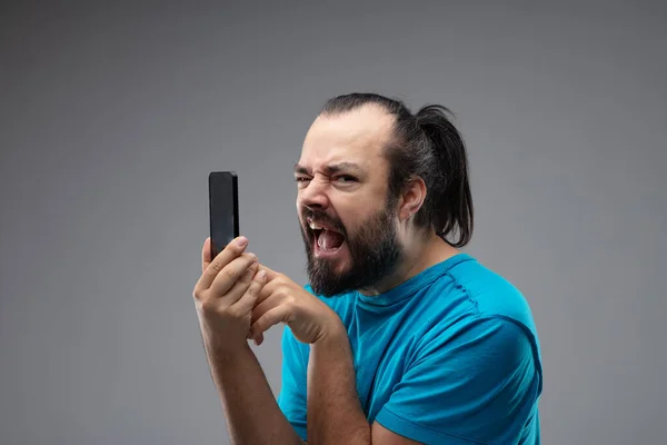 目の前にスマートフォンを持っている髭の男は 携帯電話を使用するときにカメラを見て 嘆きと泣き叫んでいます 灰色の背景に対する閉鎖的な肖像画 — ストック写真