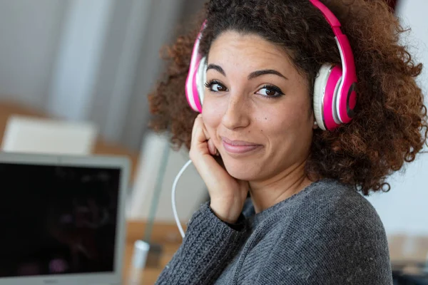 A happy, young woman with frizzy hair wearing headphones while working on a laptop from home.