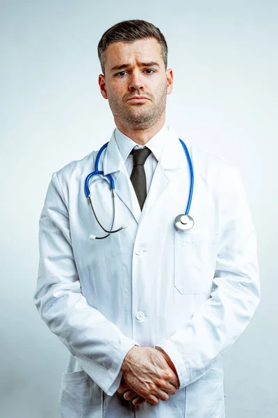 年轻的男医生 身穿白衣 头戴领带 头戴听诊器 站在那里 双手合十 满脸愁容地看着相机 背景为白色的正面肖像 — 图库照片