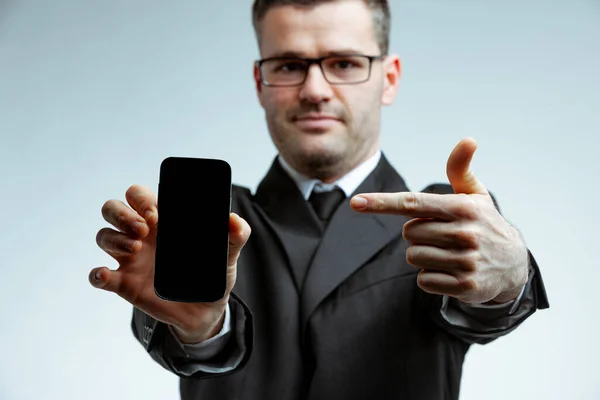男性の眼鏡とグレーのスーツのフロント選択焦点の肖像画 彼の人差し指で彼の手にスマートフォンを指して カメラに面した黒い画面で携帯電話を示しています — ストック写真