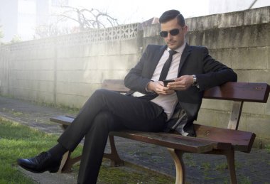 Cep telefonu kullaranarak parkta oturan takım elbiseli genç adam