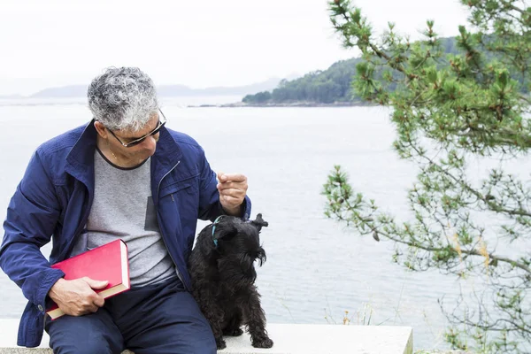 Äldre man med bok och hund sitter med seascape — Stockfoto