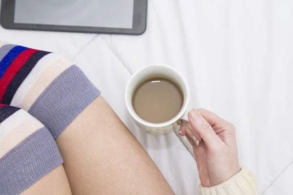 woman in socks on top of the bed watching the tablet and with a cup of coffee