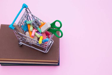 Pembe arka planda farklı kırtasiye malzemesi olan alışveriş arabası.
