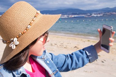 Şapkalı kız sahilde cep telefonuyla selfie çekiyor.