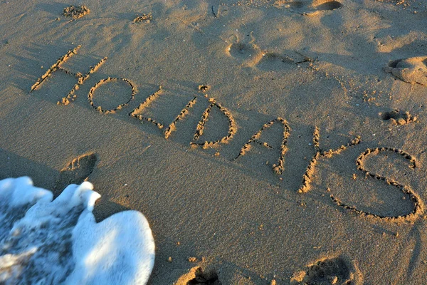 La palabra HOLIDAYS escrita en una playa de arena . —  Fotos de Stock