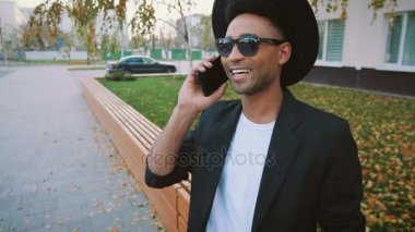 Siyah takım elbiseli ve şapkalı genç moda Afro-Amerikalı yazın sokakta telefon kullanıyor.