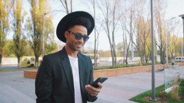 Siyah takım elbiseli ve şapkalı genç moda Afro-Amerikalı yazın sokakta telefon kullanıyor.