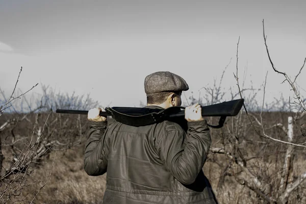 Un uomo con la schiena in giacca e jeans con un fucile da caccia in mano cammina lungo la steppa della foresta in cerca di selvaggina — Foto Stock