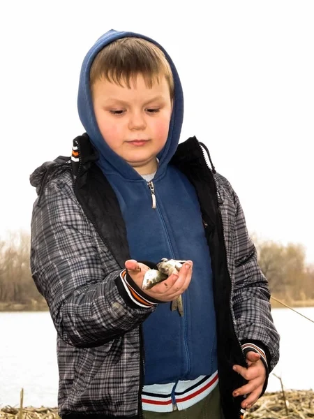 Счастливый улыбающийся мальчик стоит на берегу реки и держит в руках рыбу. Мальчик поймал рыбу. Осенняя рыбалка — стоковое фото