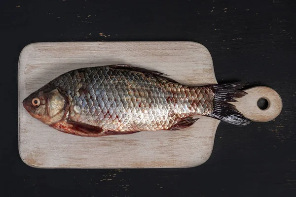 Um peixe crucian fresco encontra-se em uma placa de corte de madeira branca em um fundo de madeira preto. Vista superior — Fotografia de Stock