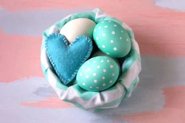 Minimal easter concept. Basket with colored easter eggs and a blue felt heart on a pink - blue wooden background