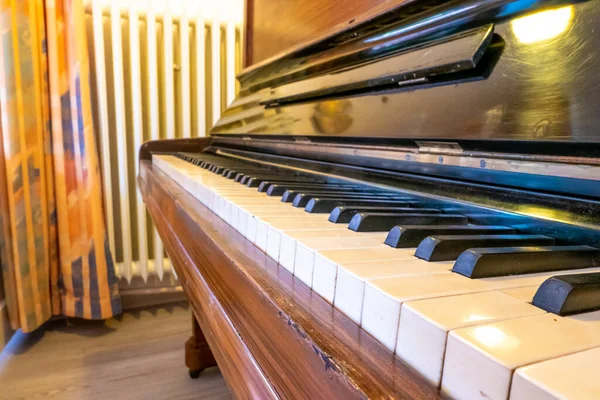 An old brown piano in a room with a heater and curtains in the background with room lighting.