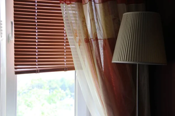 closed brown blinds, window, curtains and floor lamp with beige shade. elements of home interior on a Sunny day