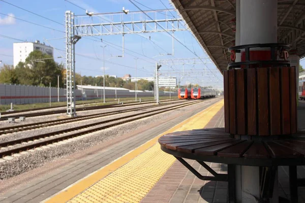 Stazione della metropolitana, panca in legno sulla piattaforma ferroviaria per una comoda attesa sullo sfondo delle rotaie e due treni in lontananza . — Foto Stock