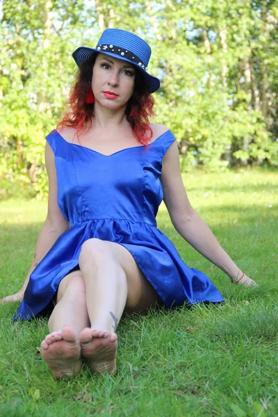 Femme dans une robe courte bleue classique et chapeau est assis étirant ses jambes vers l'avant sur la pelouse verte. cheveux roux et boucles d'oreilles — Photo
