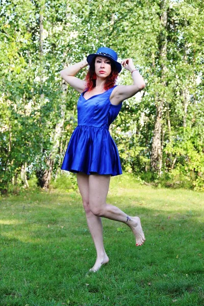 Women with red curly hair in a classic blue short dress and blue hat, jumping on one leg on the green lawn — Stock Photo, Image