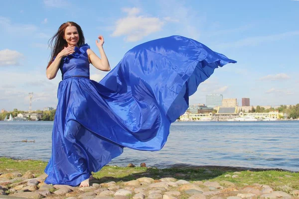 Caminar a lo largo del río. mujer en un vestido clásico azul largo que revoloteaba en el viento. reír en voz alta morena — Foto de Stock