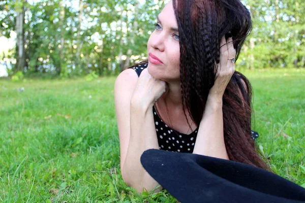 Morena melancólica se encuentra en el césped verde en el parque. mira al lado alisando el pelo largo de la mano al lado del sombrero negro — Foto de Stock