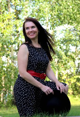 Gülümseyen esmer, uzun siyah saçlı, siyah yazlık elbiseli, kırmızı kemerli ve elinde siyah şapkası olan..