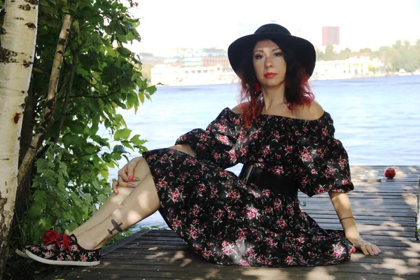 Brunette with curly red curls in a black hat and summer dress sits on a wooden bridge on the river bank on a summer day. a Cup of tea and an Apple on the background — Stock Photo, Image