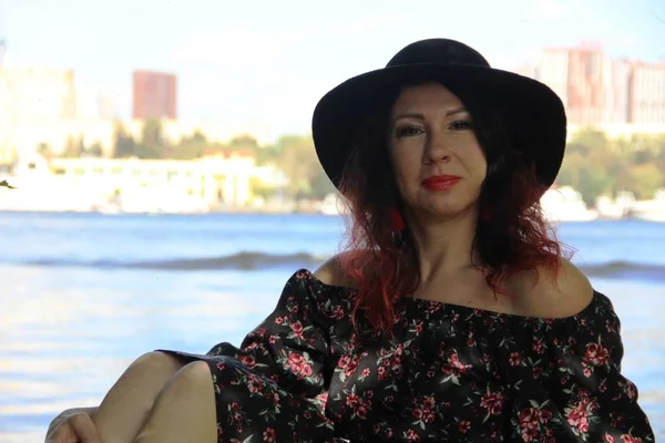 Morena con rizos rojos rizados en un sombrero negro y vestido de verano se sienta en un puente de madera en la orilla del río en un día soleado de verano, medio girado, con la mano envuelta alrededor de la rodilla — Foto de Stock