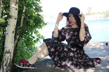 Siyah şapkalı, kıvırcık kırmızı bukleli ve yaz elbisesi olan esmer bir kadın, bir yaz günü nehir kıyısındaki ahşap bir köprüde oturur. Elleriyle şapkayı kafasında tutuyor..