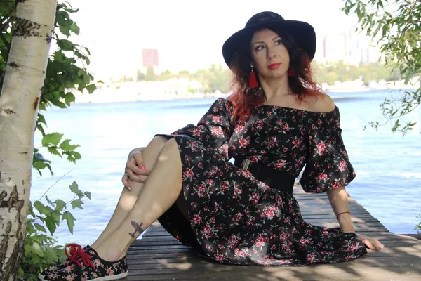 Brunette with curly red curls in a black hat and summer dress, sits on a wooden bridge on the riverbank — Stock Photo, Image