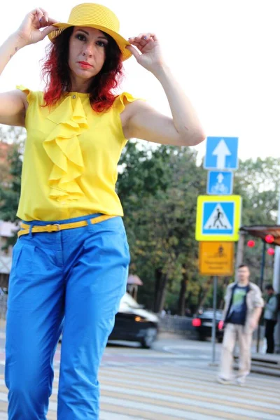 Femme aux cheveux bouclés rouges, en pantalon classique bleu, un chemisier jaune et un chapeau dans la ville, se tient sur un passage piéton, flou — Photo