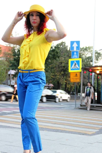 Mujer con el pelo rizado rojo, en pantalones clásicos azules, una blusa amarilla y un sombrero amarillo se encuentra en un cruce peatonal en entornos urbanos — Foto de Stock