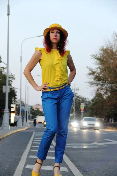 Mujer con el pelo rizado rojo, en pantalones clásicos azules, una blusa amarilla y sombrero en la carretera. noche en condiciones urbanas, luces de coche en la distancia — Foto de Stock