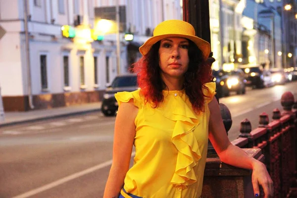 Mujer en un sombrero amarillo y ropa contra el fondo de las luces de la noche de la gran ciudad, pasando coche en la distancia. morena dama de pie en el camino . — Foto de Stock