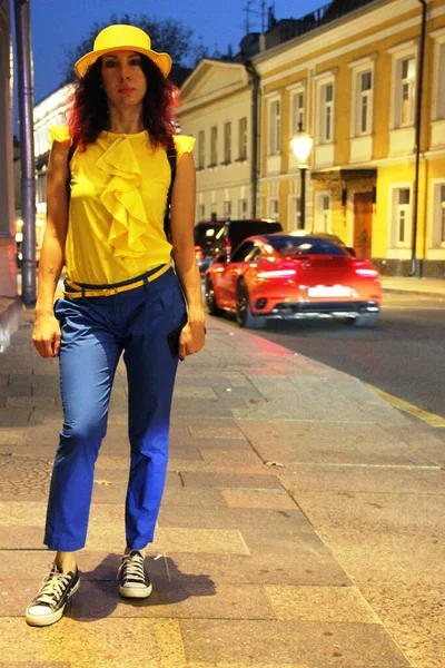 Une promenade dans la ville nocturne. femme aux cheveux bouclés rouges, chapeau jaune, pantalon bleu et baskets. costume fantaisie jaune-bleu, ville lumière du soir — Photo