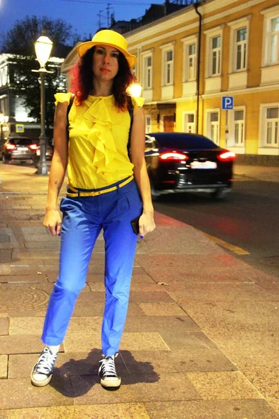 Une promenade dans la ville nocturne. Une femme aux cheveux bouclés rouges, un chapeau jaune, un pantalon bleu et des baskets. costume fantaisie jaune-bleu, lumière du soir. tonification — Photo