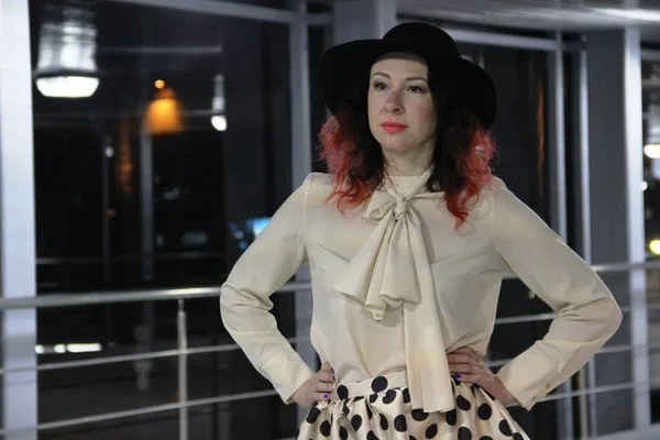 brunette in a beige blouse with a dickeys and a skirt with black polka dots. woman in a black hat with red hair against a glass wall with a railing, the night city outside the window.