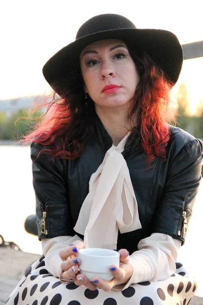 An attractive woman with red curly hair, in a black hat and with a cup of tea on the river — Stock Photo, Image