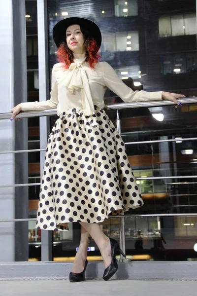 Woman in a black hat with red hair against a glass wall with a railing, the night city outside the window. brunette in a beige blouse with a dickeys and a skirt with black polka dots — Stock Photo, Image
