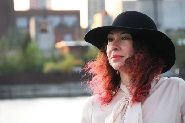 Mujer con el pelo rojo y un sombrero negro clásico junto al río de la ciudad — Foto de Stock