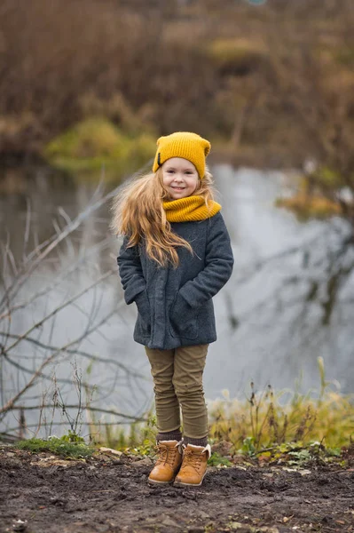 Niña con un sombrero amarillo y bufanda camina a lo largo de la orilla de un — Foto de Stock