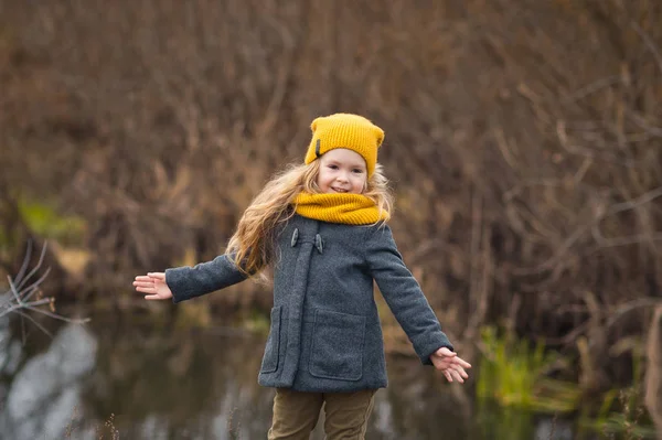 Little girl in a yellow hat and scarf walks along the shore of a — Stock Photo, Image
