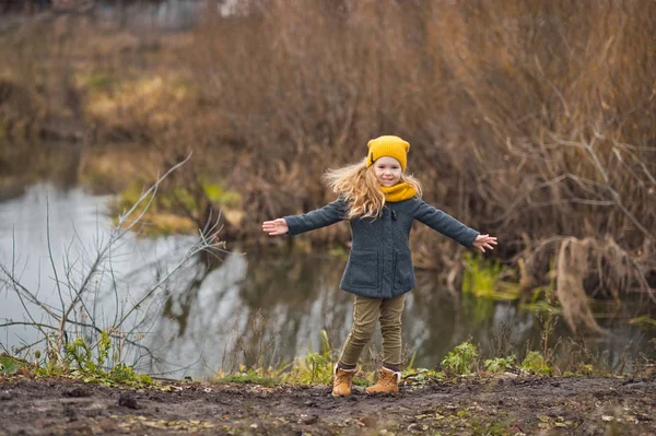 Petite fille dans un chapeau jaune et écharpe marche le long de la rive d'un — Photo