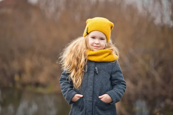Niña con un sombrero amarillo y bufanda camina a lo largo de la orilla de un — Foto de Stock