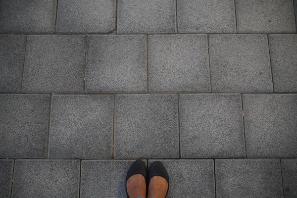 Vrouwen Zwarte casual schoenen staan en rusten op asfalt betonnen vloer met vierkante tegels. Bovenaanzicht. Betonnen vloer textuur patroon bestrating achtergrond. Selfie vrouw van voeten en benen gezien van boven. — Stockfoto