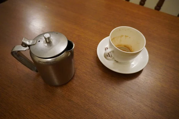 Chaleira de panela de chá de alumínio usado sujo velho com caneca de chá branco usado sujo. Chaleira de alumínio antigo com xícara de chá e saquetas de açúcar usadas vazias. Café sujo e xícara de chá meio vazio no pires na mesa. — Fotografia de Stock