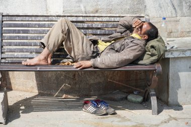 Zavallı evsiz dilenci adam ya da şehirdeki tek yönlü bir sokakta gün boyunca kirli bir tahta bankta uyuyan mülteci. Sosyal belgesel konsepti. Ayakkabılar yere. - Udaipur Hindistan: Şubat 2020
