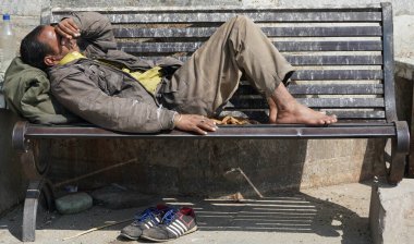 Zavallı evsiz dilenci adam ya da şehirdeki tek yönlü bir sokakta gün boyunca kirli bir tahta bankta uyuyan mülteci. Sosyal belgesel konsepti. Ayakkabılar yere. - Udaipur Hindistan: Şubat 2020